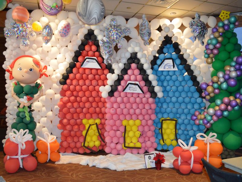 A colorful balloon display exhibited at Festival of Trees with balloons designed as an elf, presents, and houses listing sponsors of the event.