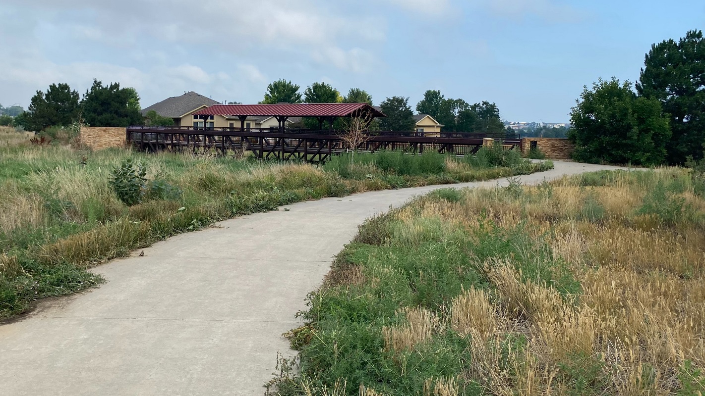 A concrete paved trail surrounded by brown grass with a red roof bridge