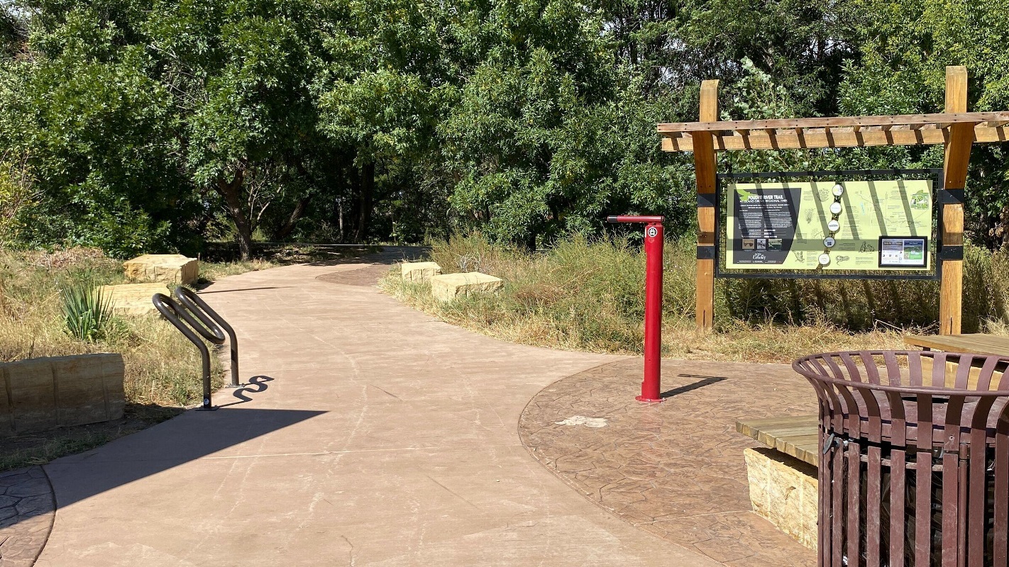 Concrete trail with red bike repair post, trees and metal bike rack.