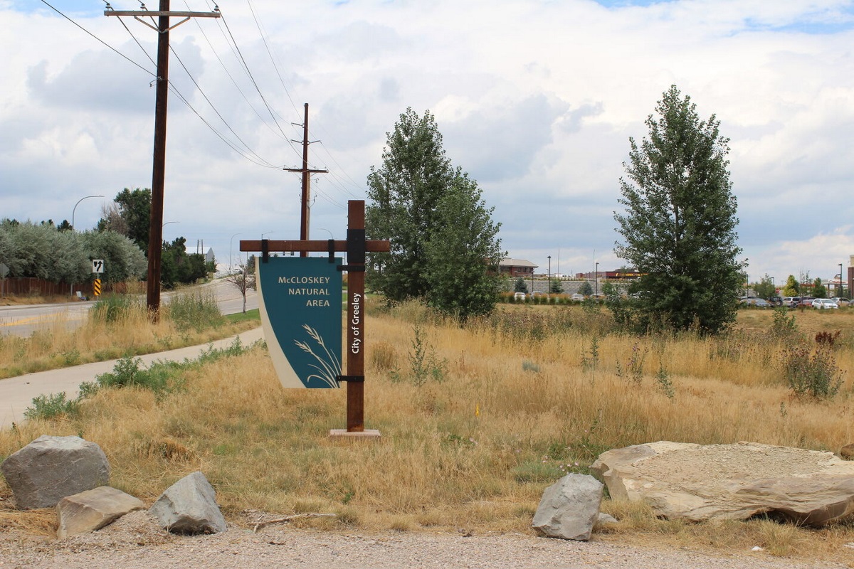 A blue sign reads &quot;Natural Area&quot; in front of grass and rocks next to a road with buildings in the background