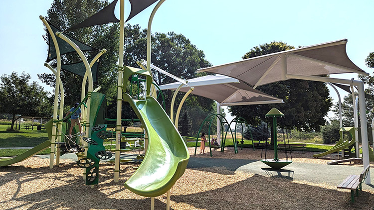 A green slide and playground with a shaded canopy, wood fiber surface, a wood bench, and tall green trees. A woman pushes a child on a swing and a little boy climbs on ropes.