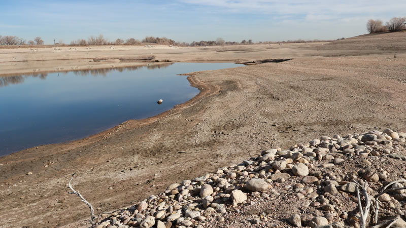 A dry, barren pond with a small patch of water remaining near the center, surrounded by sandy, rocky shores.