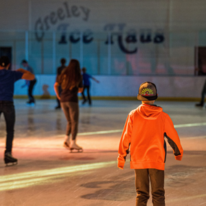 A young child in an orange shirt and backward hat skates towards others on the brightly colored Greeley Ice Haus rink