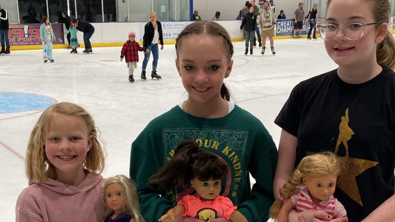 Three girls with their dolls at the Ice Haus rink during the American Girl Doll Skate, while skaters are on ice behind them.