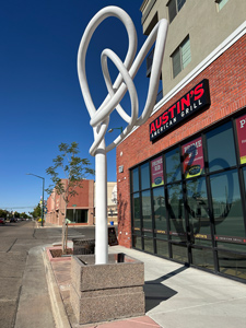 An artist's rendition of a tree using sinuous metal in curving lines, painted white against a bright blue sky in front of Greeley's Austin's restaurant.