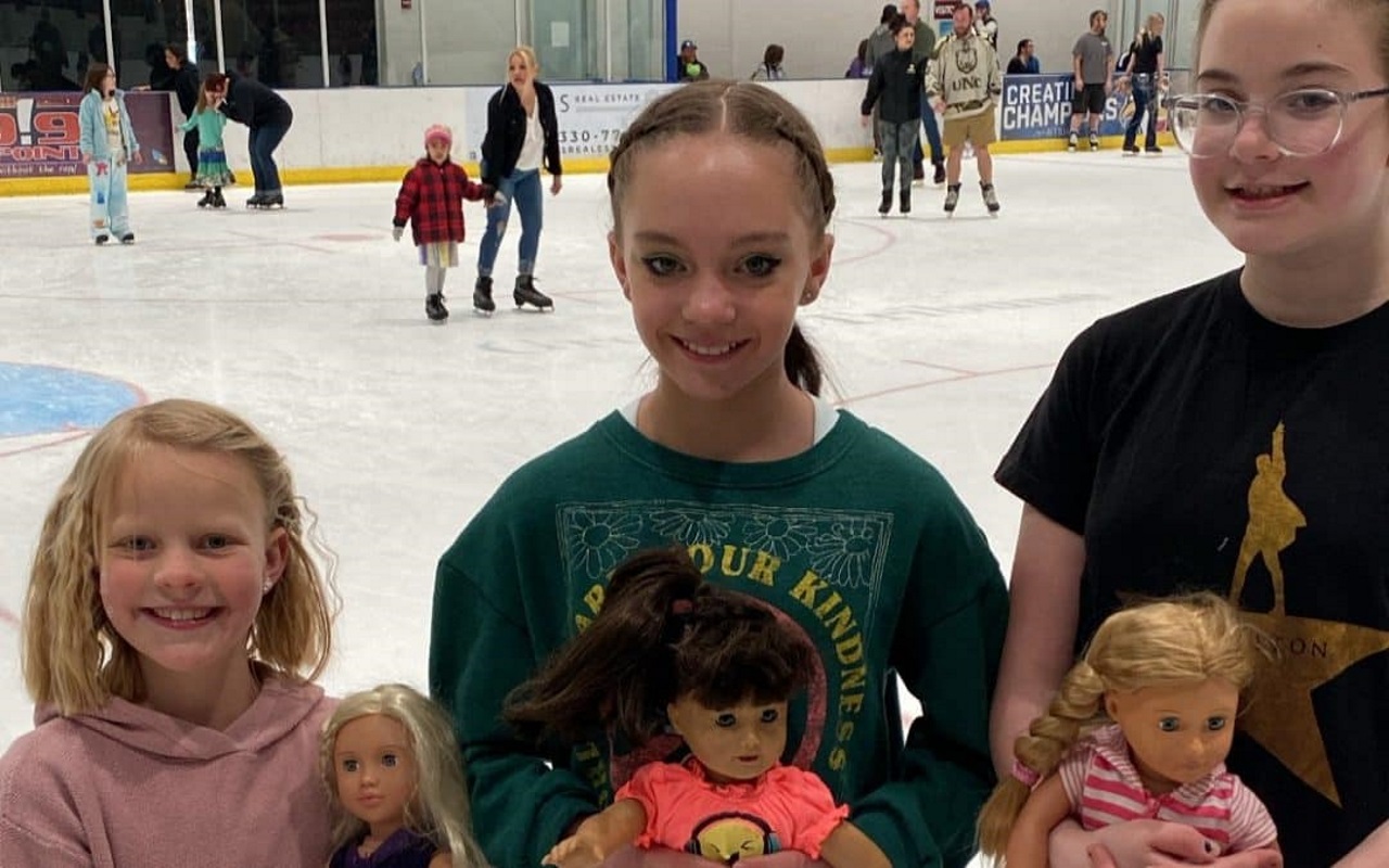 Three girls holding dolls in front of an ice rink with people skating