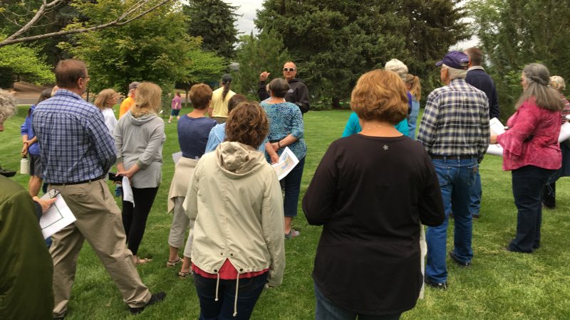 A group of people listen to a person talk about a variety of trees in the background.