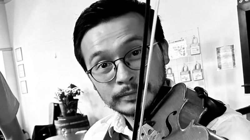 Man playing a violin looks directly at the camera. He is wearing glasses and a vest over a collared shirt. The background shows tables, chairs and some wall decorations.