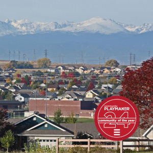 Residential area with houses and trees showing autumn colors in the foreground. Snow-capped mountains are visible in the distance under a blue sky. A circular red award badge in the bottom right reads Community Playmaker 24, community of the year.