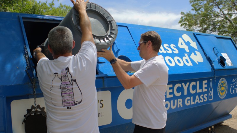 Two men in white and purple O-I t-shirts empty a trash can full of glass recyclables into a large blue dumpster labeled GLASS4GOOD.