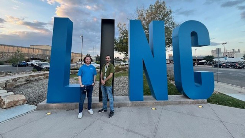 Two men stand on a sidewalk in front of an oversized sign the says LINC in large blue and black letters. There is a parking lot, buildings, and street with cars in the background.