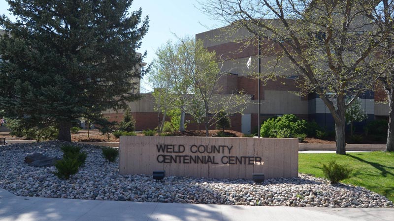 View of Weld County Centennial Center, featuring a large evergreen tree and bare trees on a landscaped area with rocks. The center’s sign is prominently displayed in the foreground with the building in the background under a clear sky.